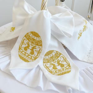 May include: White Easter-themed napkins with gold cross-stitch egg and bunny designs. The napkins are tied to a wicker basket with a large white bow. Other white linens with gold embroidery are also visible.