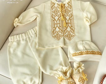 Könnte beinhalten: Ein weißes Baby-Taufoutfit mit goldener Stickerei. Das Outfit besteht aus einem langärmeligen Hemd, einer Hose, einer Mütze und Stiefeln. Das Hemd hat ein traditionelles Stickmuster auf der Vorderseite. Die Hose hat einen elastischen Bund mit Raffung. Die Mütze hat einen goldenen Besatz. Die Stiefel haben einen goldenen Quasten.