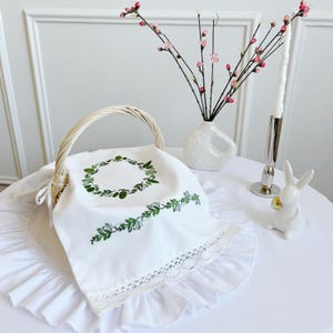 May include: A white wicker basket with a handle, containing a white cloth with green leaf embroidery and a lace trim. A white ceramic bunny figurine and a vase with pink flowers are on the table.