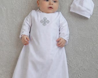 May include: A white baby baptism gown with long sleeves and a silver cross design on the chest. The gown has lace trim at the cuffs and hem. A matching white bonnet with lace trim is next to the gown.