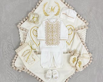 Puede incluir: Conjunto de ropa de bebé blanco con bordados dorados, que incluye una manta, gorro, camisa, pantalón y patucos. La manta presenta una "M" bordada y alas de ángel. También se incluye una pequeña bolsa con texto.