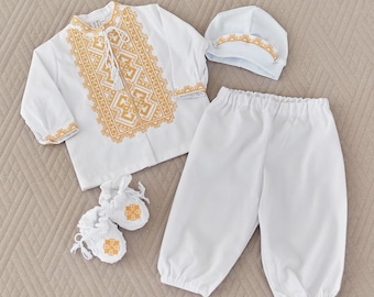 Puede incluir: Conjunto de ropa de bebé blanco con bordados dorados. El conjunto incluye una camisa de manga larga, pantalones, un gorro y patucos. La camisa presenta intrincados diseños dorados alrededor del cuello, los puños y el frente.