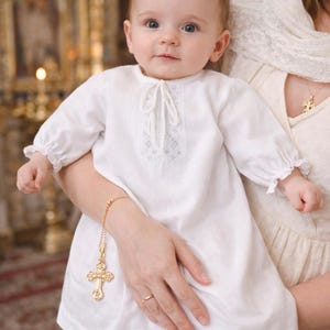 May include: A baby wearing a white baptismal gown with long sleeves and a decorative neckline. A gold cross pendant hangs from a bracelet on the baby's wrist. The baby is held by a person in a church setting.
