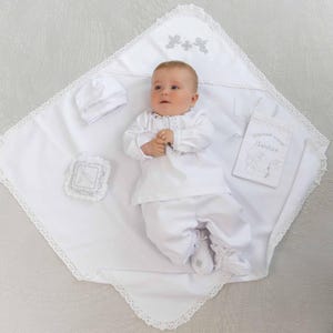 Puede incluir: Un conjunto blanco de bautizo para bebé que incluye una manta, gorro, babero y traje. La manta y el babero tienen detalles bordados y ribete de encaje. El bebé lleva el traje y los zapatos. El conjunto está diseñado para una ceremonia de bautizo.
