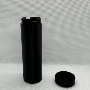 May include: A black cylindrical container with a lid. The container is made of a smooth, matte material. The lid is round and has a slightly raised edge.