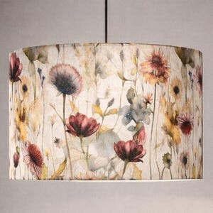 Peut inclure: Abat-jour cylindrique avec un motif floral. Le design présente des fleurs à l'aquarelle dans des tons de rouge, jaune et violet sur un fond clair. L'abat-jour est suspendu à un cordon noir et à un luminaire de plafond.