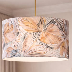 Peut inclure: Abat-jour cylindrique avec un imprimé botanique de grandes feuilles stylisées dans des tons pêche, beige et gris. Il est suspendu à une tige et un support de plafond dorés, offrant une esthétique chaleureuse et naturelle pour la décoration intérieure.