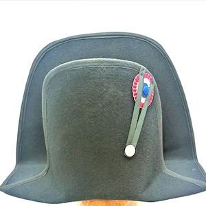 Puede incluir: Un bicorne de fieltro verde oscuro con una escarapela roja, blanca y azul. El sombrero tiene un ala ancha y una corona alta.