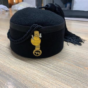 Könnte beinhalten: Ein schwarzer Filzfez mit goldener Quaste und einem goldenen Emblem mit einer Säule und der Zahl "74".