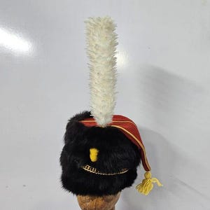 May include: A black fur hat with a red and gold trim and a white feather plume. The hat has a gold chain detail around the brim.