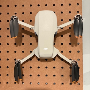 Może przedstawiać: Biały dron z czterema czarnymi śmigłami i pomarańczowymi akcentami jest zamontowany na ścianie z płyty perforowanej. Dron ma logo DJI na spodzie.