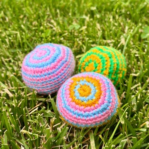 Puede incluir: Tres bolas de ganchillo coloridas descansan en hierba verde. Las bolas presentan círculos concéntricos de hilo rosa, azul, amarillo y verde. Las bolas parecen hechas a mano y son un objeto decorativo y lúdico.