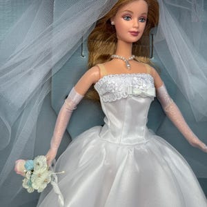 Peut inclure: Poupée de mariée vêtue d'une robe de mariée en satin blanc, d'un voile et de gants. La poupée a de longs cheveux blonds et tient un petit bouquet de fleurs. Un collier et des boucles d'oreilles en perles complètent le look.