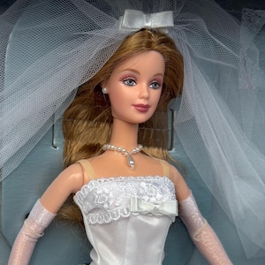 May include: A doll with long brown hair wearing a white strapless gown with lace trim and a small bow. The doll features a white tulle veil with a bow, a pearl-style necklace with a teardrop pendant, small pearl stud earrings, and sheer white gloves.