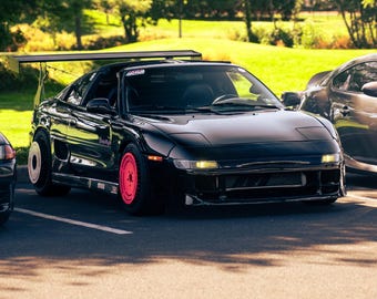 May include: A black sports car featuring a large rear wing, a front bumper with integrated lights, and mismatched wheels with a bright pink front wheel and a white rear wheel cover. The vehicle has a low-profile body and a sleek, aerodynamic design.