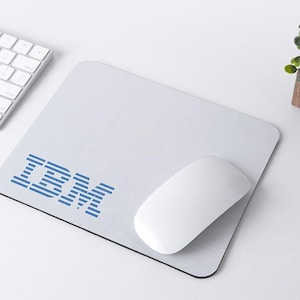 Può includere: Un tappetino per mouse bianco con un logo IBM blu stampato su di esso. Un mouse per computer bianco è sul tappetino per mouse.
