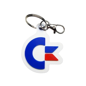 Può includere: Portachiavi bianco con il logo Commodore in blu e rosso. Il logo è una forma stilizzata a "C" con un elemento a forma di bandiera. Il portachiavi ha un anello e un moschettone argentati, adatti per chiavi o borse.
