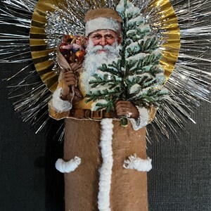 May include: A vintage Christmas ornament featuring a Santa Claus figure holding a small tree and a bundle of toys. The ornament is made of paper mache and has a brown felt body with white fur trim. The Santa Claus figure is surrounded by a gold and silver tinsel halo.