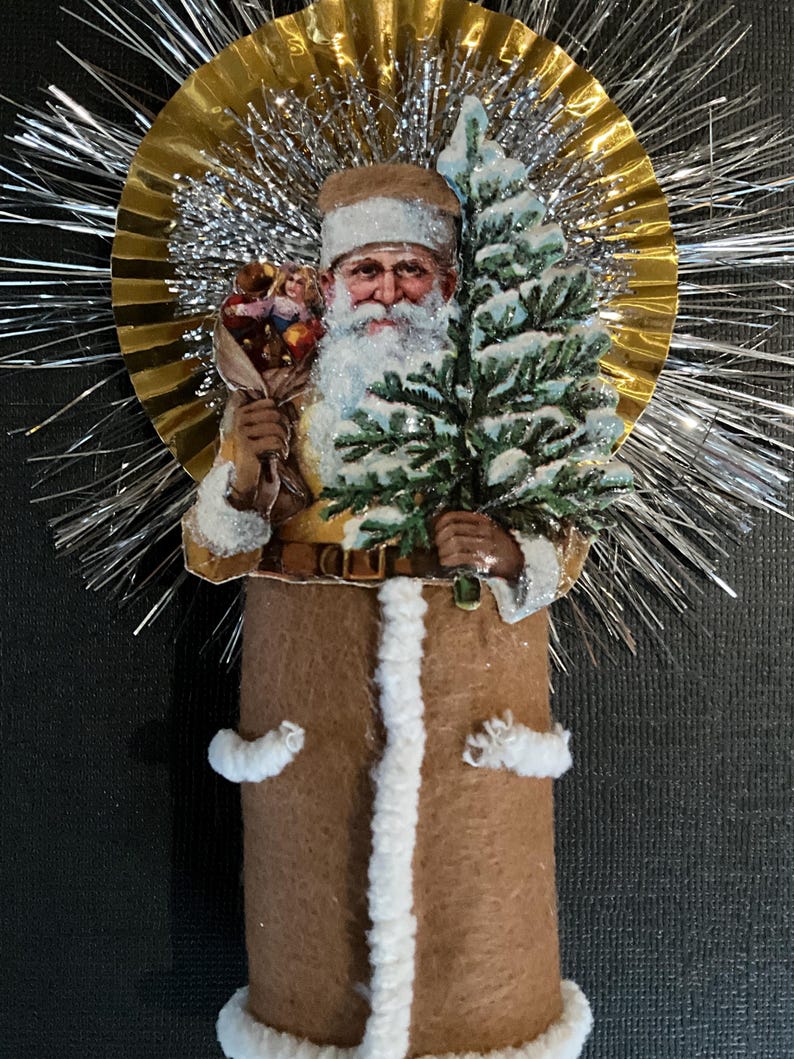 May include: A vintage Christmas ornament featuring a Santa Claus figure holding a small tree and a bundle of toys. The ornament is made of paper mache and has a brown felt body with white fur trim. The Santa Claus figure is surrounded by a gold and silver tinsel halo.
