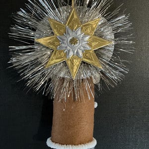 May include: A brown felt angel ornament with a white fur trim and white paper legs. The angel is topped with a silver and gold star with a silver and gold flower design.