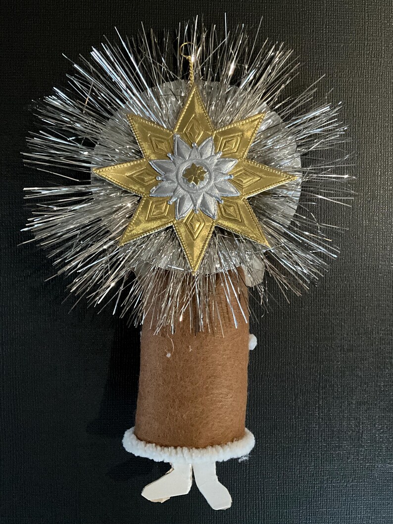 May include: A brown felt angel ornament with a white fur trim and white paper legs. The angel is topped with a silver and gold star with a silver and gold flower design.