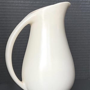 May include: Cream-coloured ceramic jug with a curved handle and a spout. The jug has a rounded body and a smooth, glossy finish. The handle is large and easy to grip. The jug is likely used for serving beverages.