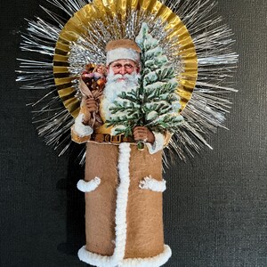 May include: A vintage-style Christmas ornament featuring a Santa Claus figure holding a small Christmas tree. The ornament is made of paper and fabric and has a gold and silver tinsel halo.
