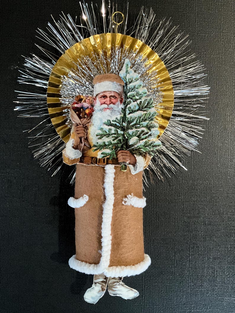 May include: A vintage-style Christmas ornament featuring a Santa Claus figure holding a small Christmas tree. The ornament is made of paper and fabric and has a gold and silver tinsel halo.