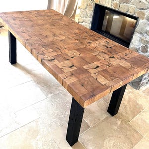 Può includere: Un tavolo da pranzo rettangolare con un piano spesso costruito con numerosi piccoli pezzi di legno marrone naturale tagliati a forma quadrata, con venature e nodi scuri visibili, sostenuto da quattro robuste gambe verticali nere.
