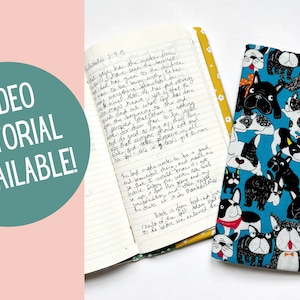 May include: A blue fabric journal cover with a black and white dog pattern. The cover is closed and has a yellow ribbon bookmark. The text "VIDEO TUTORIAL AVAILABLE!" is in a teal circle on a pink background.