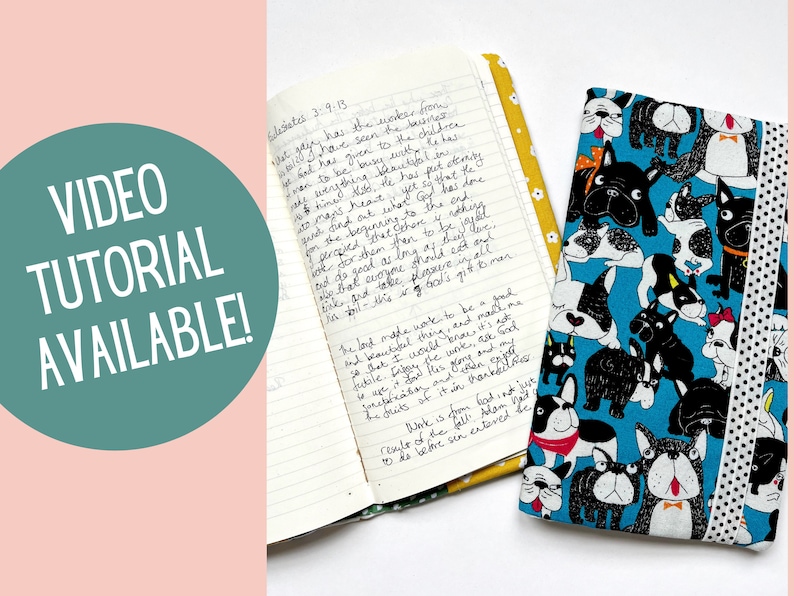 May include: A blue fabric journal cover with a black and white dog pattern. The cover is closed and has a yellow ribbon bookmark. The text "VIDEO TUTORIAL AVAILABLE!" is in a teal circle on a pink background.