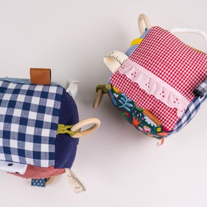 May include: Two colorful fabric cubes with different textures and patterns. One cube has a blue and white gingham pattern, a brown leather tag, and a wooden ring. The other cube has a red and white gingham pattern, a pink lace trim, and a wooden ring.