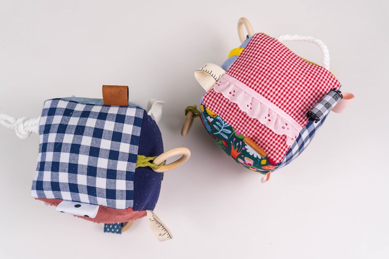 May include: Two colorful fabric cubes with different textures and patterns. One cube has a blue and white gingham pattern, a brown leather tag, and a wooden ring. The other cube has a red and white gingham pattern, a pink lace trim, and a wooden ring.