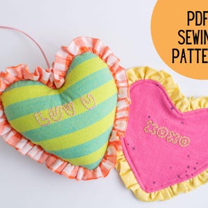 Op de afbeelding: Twee hartvormige kussens met gekrulde randen. Eén is groen en wit gestreept met het woord "Love" geborduurd erop. De andere is roze met het woord "XOXO" geborduurd erop.  PDF-naai-patroon.