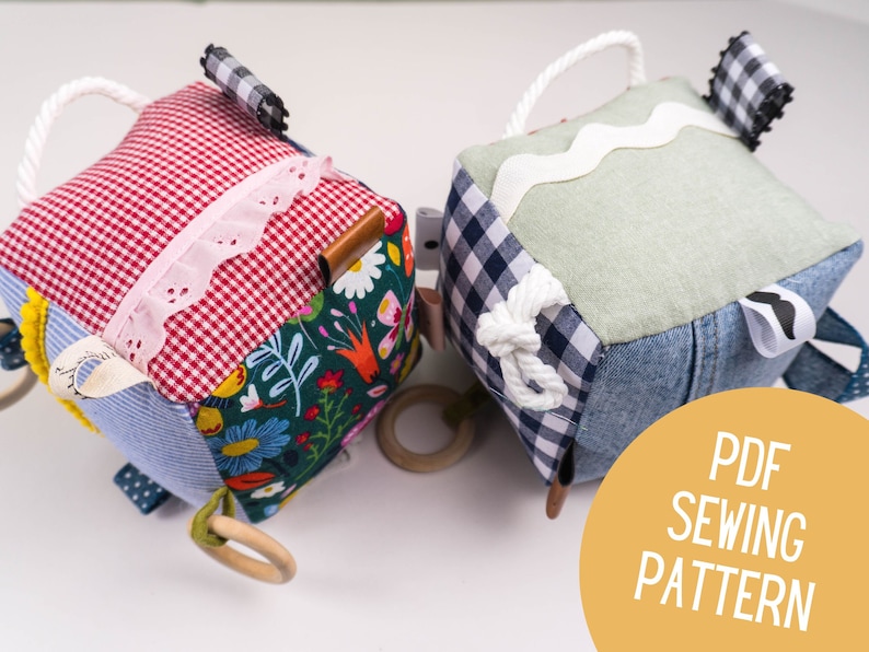 May include: Two handmade fabric baby sensory cubes with different textures and colors. The cubes have a variety of features, including ribbons, rings, and fabric scraps. The cubes are perfect for babies to explore and learn about different textures and colors. PDF Sewing Pattern.