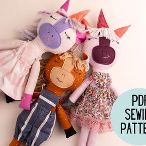 May include: Three handmade fabric dolls: a unicorn with a purple mane and pink dress, a horse with an orange mane and blue overalls, and a pink doll with a floral dress. The dolls are soft and cuddly, perfect for children. Text on the image: PDF SEWING PATTERN.