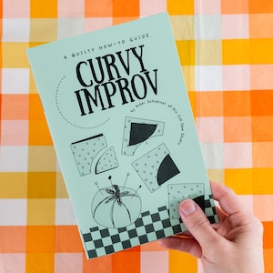 May include: A teal blue guide book titled "Curvy Improv: A Quilty How-To Guide" by Nikki Schreiner of Pin-Cut Sew Studio. The book features illustrations of curved fabric pieces and sewing pins.
