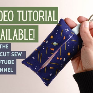 Pode incluir: Uma pequena bolsa de tecido azul com fecho de correr e um porta-chaves. A bolsa tem um padr&atilde;o colorido de estrelas, planetas e formas geom&eacute;tricas. O texto "VIDEO TUTORIAL AVAILABLE! ON THE PIN CUT SEW YOUTUBE CHANNEL" &eacute; vis&iacute;vel no lado esquerdo da imagem.