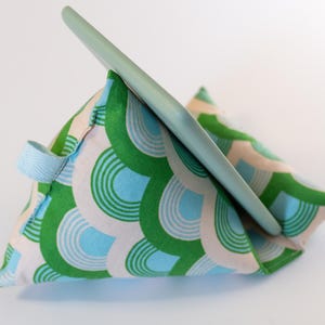 May include: A green, blue, and white patterned fabric pillow stand for a tablet or phone. The stand is shaped like a triangle and has a handle on the side.