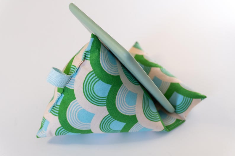 May include: A green, blue, and white patterned fabric pillow stand for a tablet or phone. The stand is shaped like a triangle and has a handle on the side.