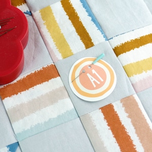 May include: A close-up of a fabric patchwork quilt with a circular label that says "A1" in orange text. The label is pinned to the quilt with a blue pin. The quilt is made of different fabrics with stripes of orange, yellow, and blue.