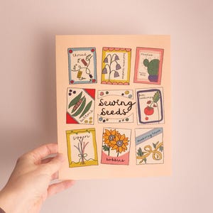 May include: A hand holds a print with nine illustrated seed packets. The packets are in colourful frames, with images of thread, flowers, a cactus, beans, a tomato, sunflowers, and scissors. The words "Sewing Seeds" are in the centre.