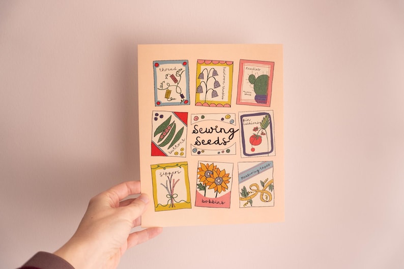 May include: A hand holds a print with nine illustrated seed packets. The packets are in colourful frames, with images of thread, flowers, a cactus, beans, a tomato, sunflowers, and scissors. The words "Sewing Seeds" are in the centre.