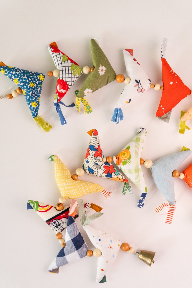 May include: A colorful garland of fabric birds with wooden beads and a bell. The birds are made of different patterned fabrics, including floral, plaid, and polka dot.