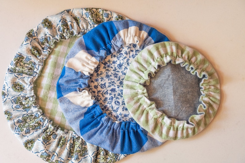 May include: Three round fabric covers in different sizes and patterns. The largest cover is blue and white with a floral pattern, the middle cover is green and white with a checkered pattern, and the smallest cover is blue and white with a floral pattern.
