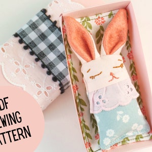 May include: A PDF sewing pattern for a small, white bunny with pink ears and a light blue dress. The bunny is lying in a pink box with a floral print lining.