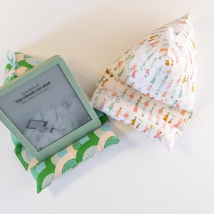 May include: A green and white patterned pillow with a  Kindle e-reader on top. The e-reader screen shows the text "Up to 90% off Top ebooks on deal". A second pillow with a white background and a pattern of colorful people is also shown.