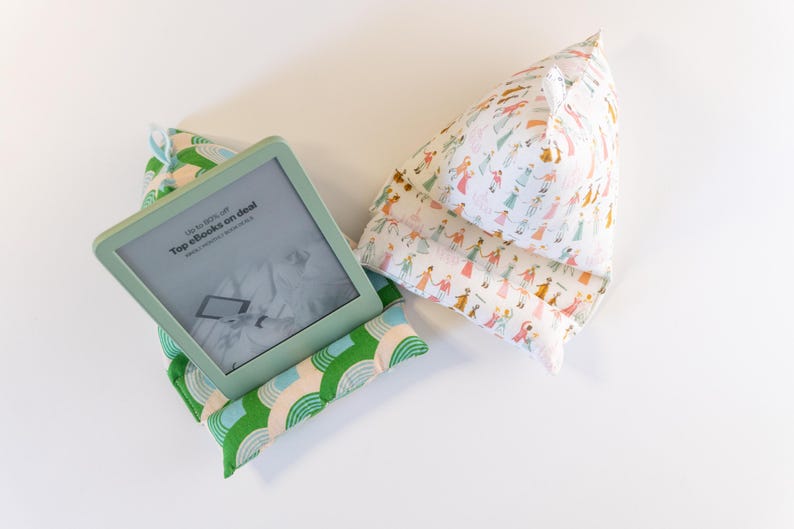 May include: A green and white patterned pillow with a  Kindle e-reader on top. The e-reader screen shows the text "Up to 90% off Top ebooks on deal". A second pillow with a white background and a pattern of colorful people is also shown.
