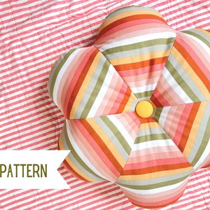 May include: A flower-shaped pillow with colorful striped fabric in shades of pink, orange, green, and white. The pillow has a yellow button in the center and rests on a pink and white striped surface. The text "PDF PATTERN" is visible.
