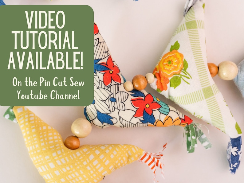 May include: A garland of colorful fabric triangles with wooden beads strung between them. The fabric is patterned with flowers and stripes. The garland is a DIY project with a video tutorial available on the Pin Cut Sew YouTube channel.